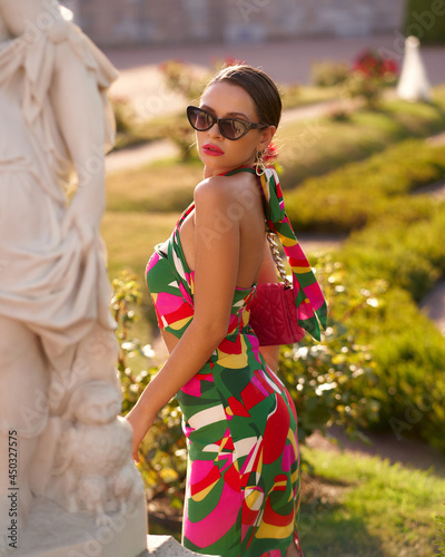 Young elegant fashionable woman wearing colorful dress, sunglasses and holding pink handbag posing in beautiful garden on a sunny summer day photo