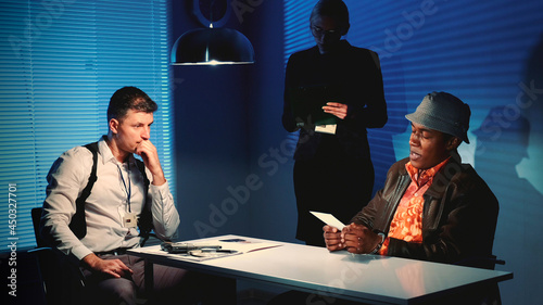 Medium shot of Detective showing picture of suspected robber's accomplice during interview in police office. In the background there is a female detective writing notes of the interrogation. photo