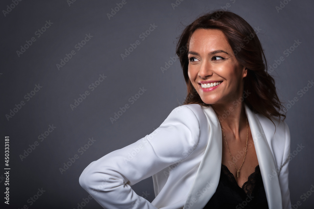 Studio portrait shot of attractive middle aged woman with toothy smile wearing white blazer while standing at isolated dark grey background. Copy space. Studio portrait shot of attractive middle aged woman with toothy smile wearing white blazer while standing at isolated dark grey background. Copy space.