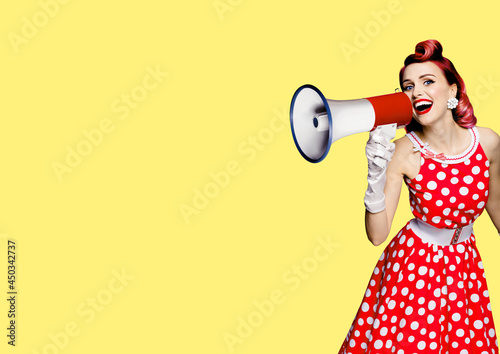 Portrait of red purple haired woman holding mega phone, shout advertising something. Girl in pin up style dress. Isolated on yellow color background with mock up. Female model in retro fashion concept