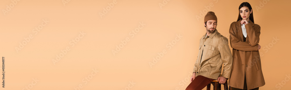 trendy man in beanie sitting near stylish woman holding hand near face isolated on beige, banner trendy man in beanie sitting near stylish woman holding hand near face isolated on beige, banner