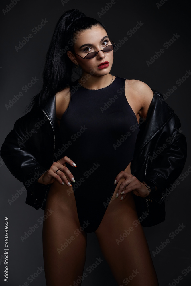 beautiful woman with a sports figure in a black bodysuit, black jacket and sunglasses posing in the studio on a gray background beautiful woman with a sports figure in a black bodysuit, black jacket and sunglasses posing in the studio on a gray background