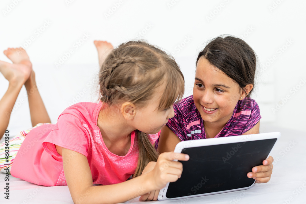 Two cute little girls are using a digital tablet and smiling while lying on bed in children's room Two cute little girls are using a digital tablet and smiling while lying on bed in children's room