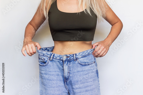 Cropped shot of a young slim woman dressed in wide jeans and short top demonstrates successful weight loss isolated on a light color gray background. Diet, wellness and health nutrition concept