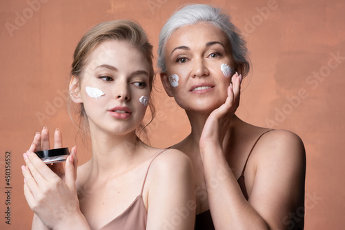  Two female generations mature mother, young adult daughter with jar applying moisturizing sunscreen cream on skin together. 