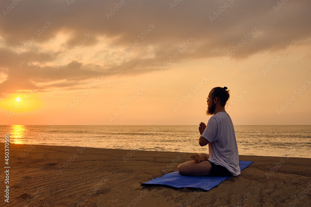 Man practices yoga meditation sitting in padmasana during sunrise. Man sits in lotus position at beach opposite rising sun and its reflection in sea. Portrait of man meditating at beach Man practices yoga meditation sitting in padmasana during sunrise. Man sits in lotus position at beach opposite rising sun and its reflection in sea. Portrait of man meditating at beach
