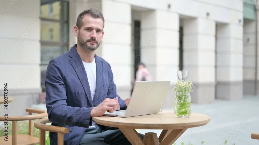 Man Looking at Camera While using Laptop, Sitting in Outdoor Cafe Man Looking at Camera While using Laptop, Sitting in Outdoor Cafe