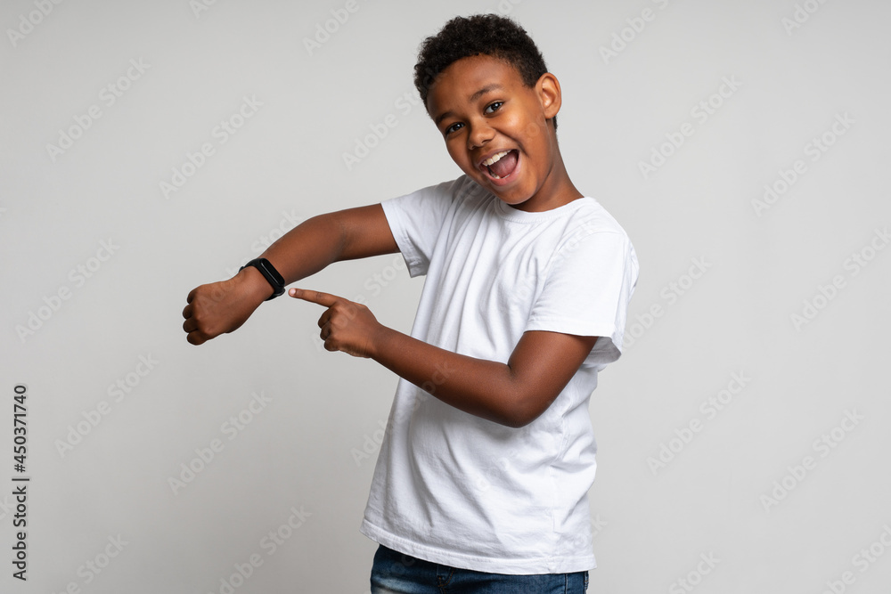 Cheerful positive boy in white shirt pointing at wristwatch on hand and smiling, showing new smart clock, checking indicators. Indoor studio shot isolated on white background Cheerful positive boy in white shirt pointing at wristwatch on hand and smiling, showing new smart clock, checking indicators. Indoor studio shot isolated on white background