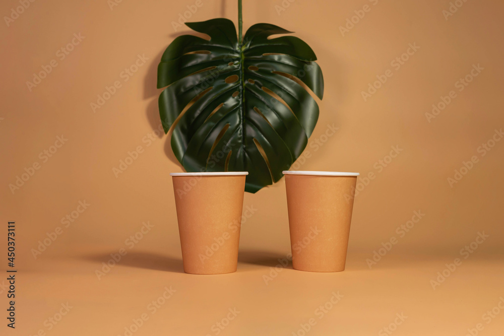 Fototapeta premium Monstera sheet, paper disposable eco-friendly cups on beige isolated background. Eco-concept.