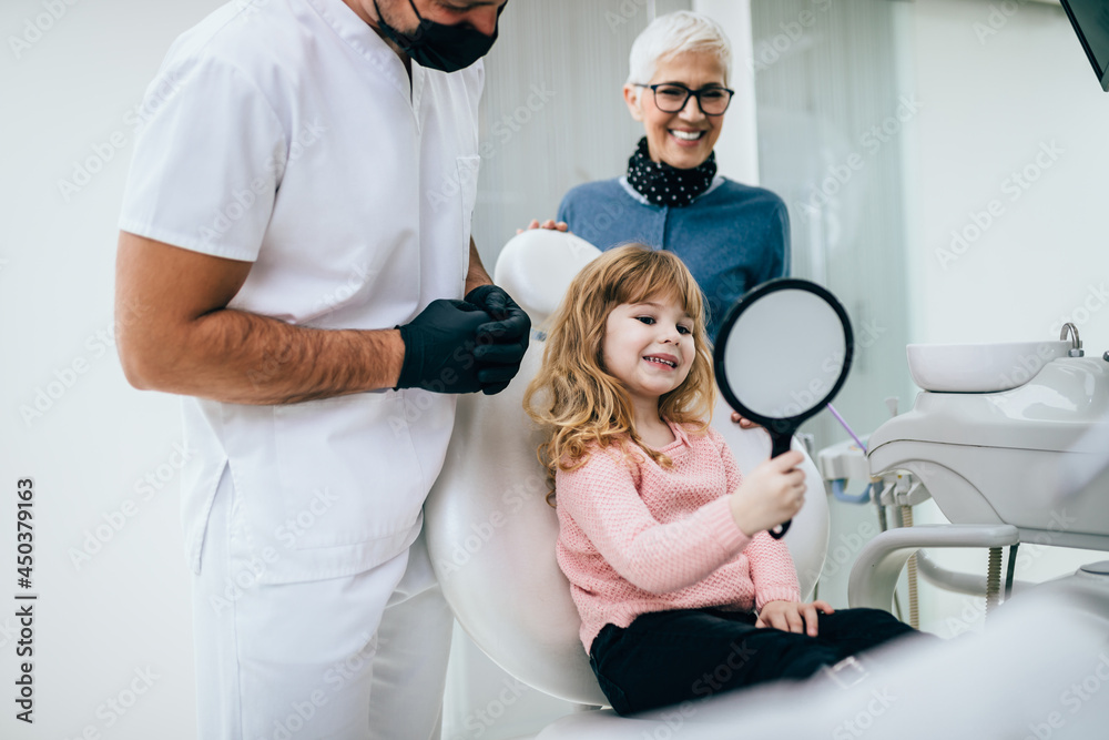 Obraz premium Cute little girl sitting on dental chair and having dental treatment. Her grandmother is with her. Dentist is wearing protective face mask due to coronavirus pandemic.