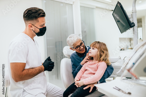 Cute little girl sitting on dental chair and having dental treatment. Her grandmother is with her. Dentist is wearing protective face mask due to coronavirus pandemic.