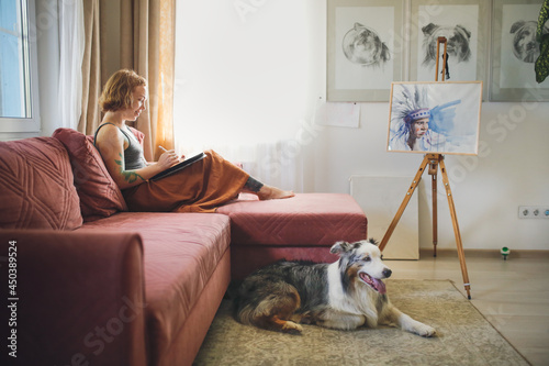cute red-haired girl artist with freckles draws on tablet in cozy living room with an Australian shepherd dog, in real interior with dog sitting floor near sofa, lifestyle