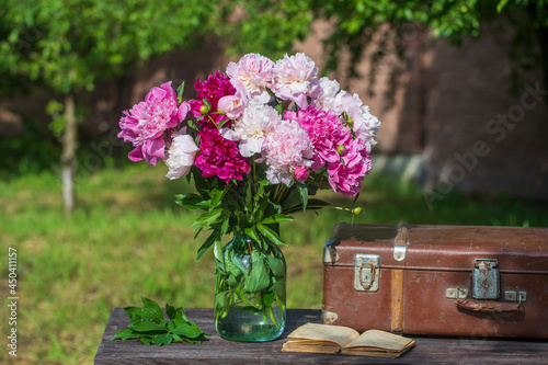 Beautiful bouquet of flowers with red, pink and white peonies in a glass jar with water and old suitcase, book on a wooden table in garden
