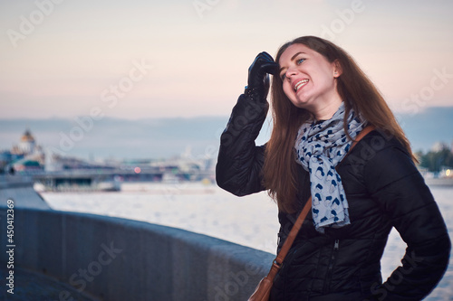 A woman in black jacket and blue scarf standing on a river embankment in the city © mediaeugene