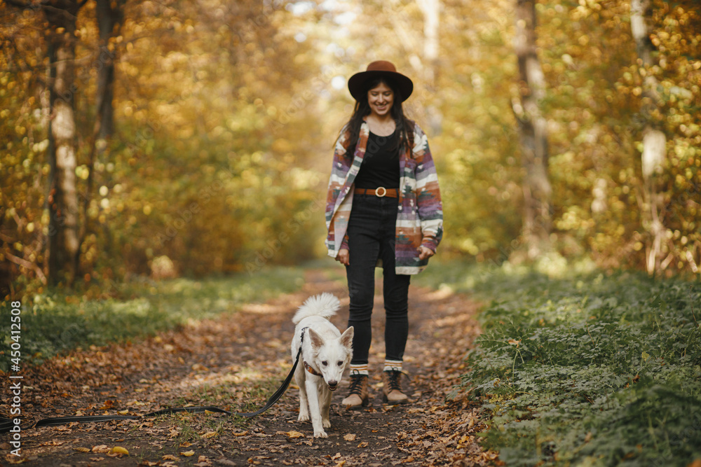 Fototapeta premium Cute dog walking on background of stylish woman traveler owner in sunny autumn woods. Traveling with pet, loyal companion. Young female hipster hiking with swiss shepherd white dog.