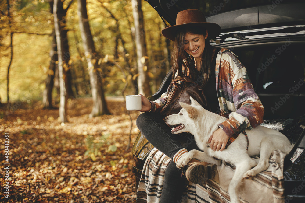 Stylish hipster woman with cup and backpack sitting with cute dog in car trunk in sunny autumn woods. Travel and road trip with pet. Space for text. Young female traveler hugging sweet white dog - obrazy, fototapety, plakaty 