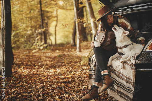 Stylish woman traveler kissing cute dog in car trunk in sunny autumn woods. Travel and road trip with pet. Space for text. Young hipster female travelling with sweet white dog, loyal companion