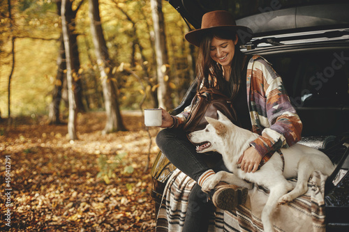 Stylish hipster woman with cup and backpack sitting with cute dog in car trunk in sunny autumn woods. Travel and road trip with pet. Space for text. Young female traveler hugging sweet white dog