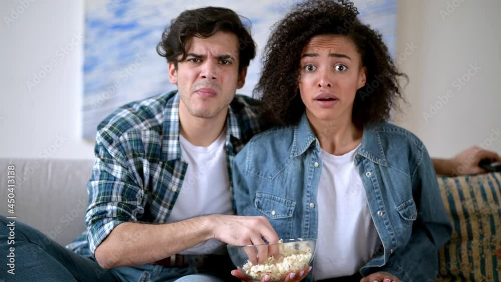 Mixed race family couple sitting at home on sofa in casual clothes, watching tv show, exciting movie or football match, eating popcorn, worried, spending weekend together at home, emotional faces Mixed race family couple sitting at home on sofa in casual clothes, watching tv show, exciting movie or football match, eating popcorn, worried, spending weekend together at home, emotional faces
