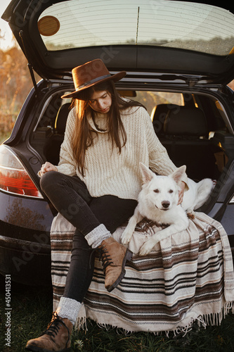 Stylish woman in hat and sweater sitting and hugging cute dog in car trunk in evening autumn field. Road trip with pet.Young hipster female travelling with sweet white dog. Travel