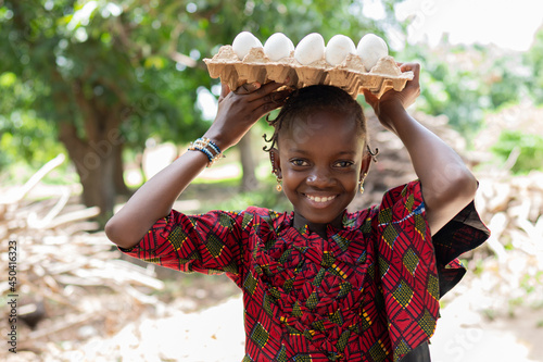 Pretty little African girl with earrings and bangles in a sophisticatedly cut dress, posing with a carton full of eggs on her head, smiling into the camera