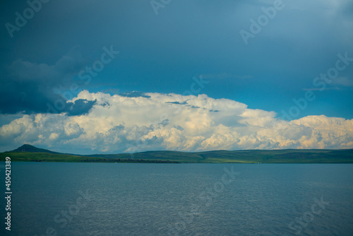 The opposite shore of the azure lake against the blue sky with white clouds. © Vectorina