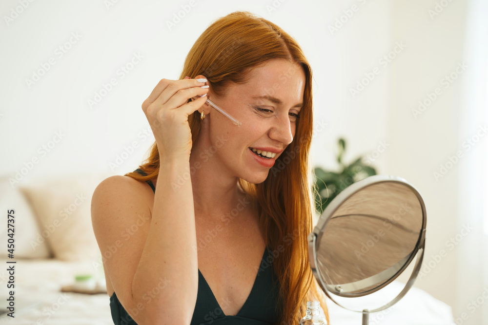 Enjoyable red hair young girl doing beauty routine with moisturizing face serum at home. Redhead smiley beautiful woman using facial natural cosmetic and looking to the mirror.