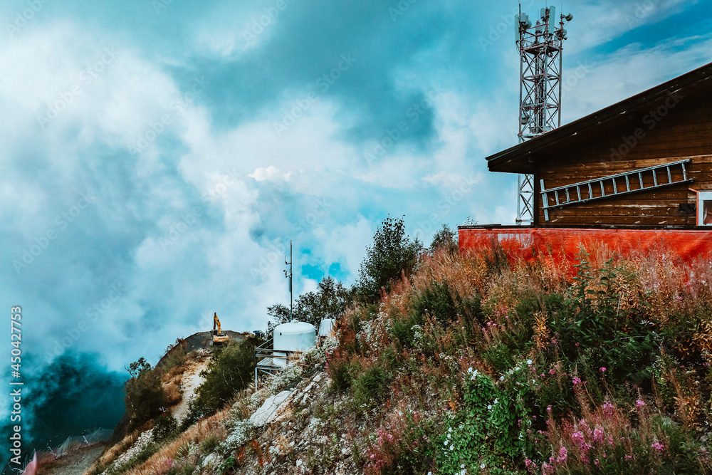 Fototapeta premium Secluded farmhouse on edge of mountain with panoramic view. Ladder on wall and telecommunication equipment on cell phone tower, communication antenna. Green plants on rocks. Tranquil scene in Europe.