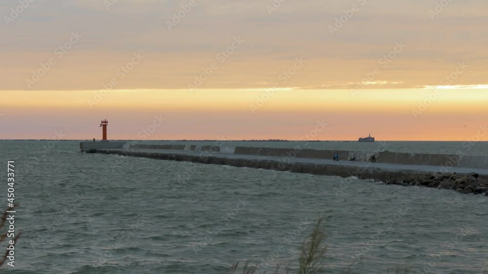 Beautiful scenic view of pink sunset with silhouettes of people walking on a stone port pier, sun rays shining trough the fast moving clouds, Karosta, Liepaja, wide low angle shot Beautiful scenic view of pink sunset with silhouettes of people walking on a stone port pier, sun rays shining trough the fast moving clouds, Karosta, Liepaja, wide low angle shot