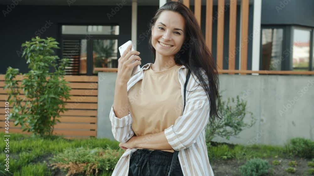 Slow motion portrait of cheerful young brunette standing outdoors holding smart phone smiling looking at camera on warm summer day. People and gadgets concept. Slow motion portrait of cheerful young brunette standing outdoors holding smart phone smiling looking at camera on warm summer day. People and gadgets concept.