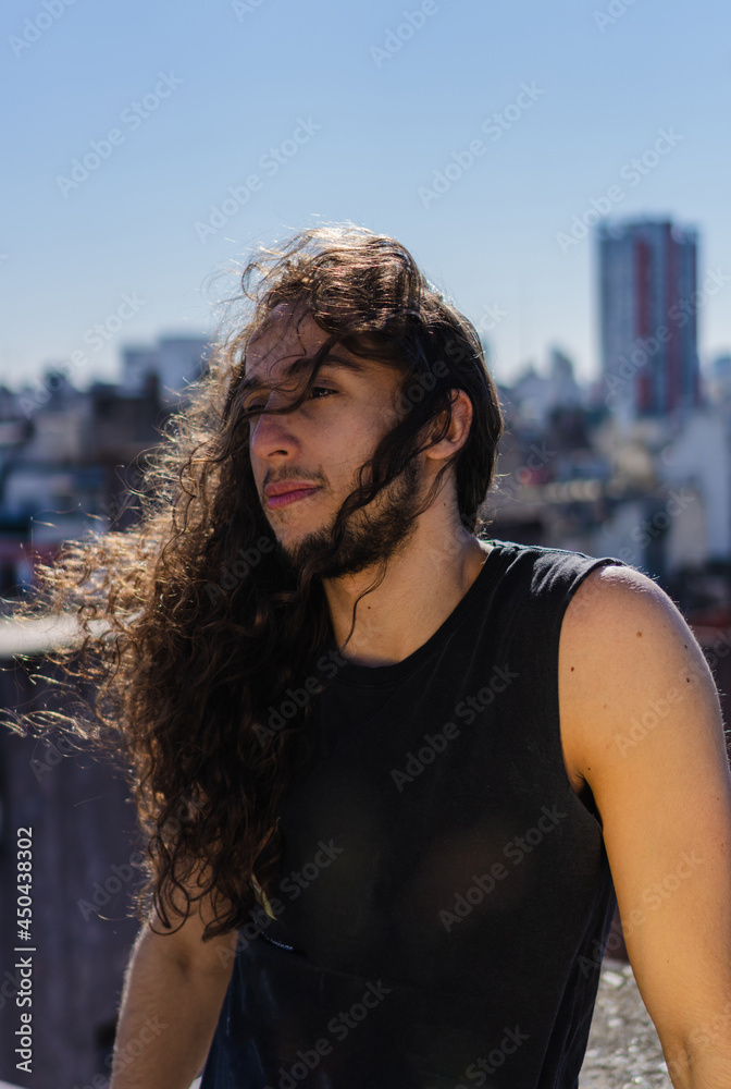 young brazilian man with long hair and blond curls against the light, blue sky background, looking away, vertical picture young brazilian man with long hair and blond curls against the light, blue sky background, looking away, vertical picture