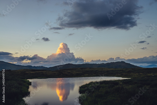 Scenic sunset landscape with yellow big cloud in form of explosion above mountain lake. Awesome giant cloud reflected in lake water in sunrise. Huge cloud of illuminating color in dawn gradient sky.