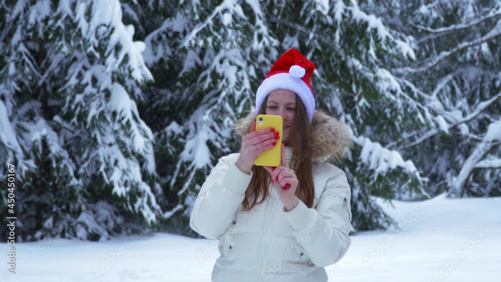 Woman with phone in winter forest. Snowdrifts and snow on spruce trees. Santaclaus hat young girl. fun before Christmas walk. take photo and video call. blogger writes a vlog. Smile at smartphone. Woman with phone in winter forest. Snowdrifts and snow on spruce trees. Santaclaus hat young girl. fun before Christmas walk. take photo and video call. blogger writes a vlog. Smile at smartphone.