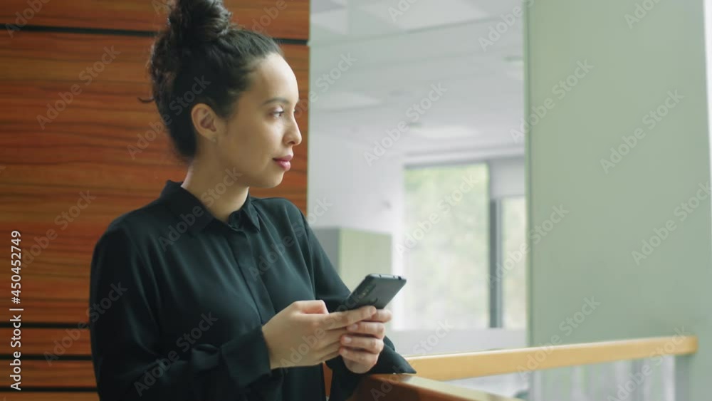 Beautiful mixed race businesswoman standing by railing in office, typing on smartphone and smiling Beautiful mixed race businesswoman standing by railing in office, typing on smartphone and smiling