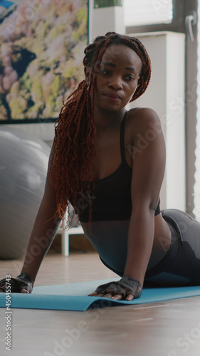 Flexible black woman practicing sport during yoga morning workout sitting on fitness map in living room. Adult stretching body muscles watching training aerobic video using laptop computer