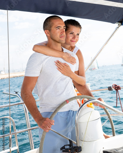 Young loving couple standing at yacht wheel enjoying sea trip along coast of Barcelona on sunny day © JackF