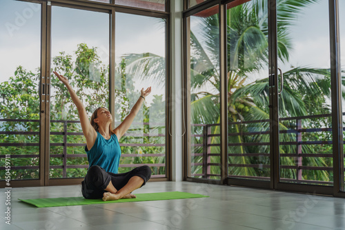 An attractive 40-year-old middle-aged woman practices yoga in a panoramic window room overlooking the garden at dawn in the sunshine. Meditation mindfulness healthy lifestyle concept.