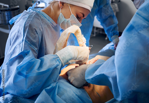 Doctors in blue uniforms performing surgical operation in operating room. Plastic surgeon and assistants removing excess fat from patient abdomen. Concept of abdominoplasty, cosmetic plastic surgery.