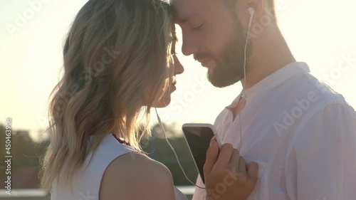 Woman and man are standing on roof together and hugging listening to music with headphones using playlist in smartphone. Close-up. Contour light. meeting of young lovers on roof in evening