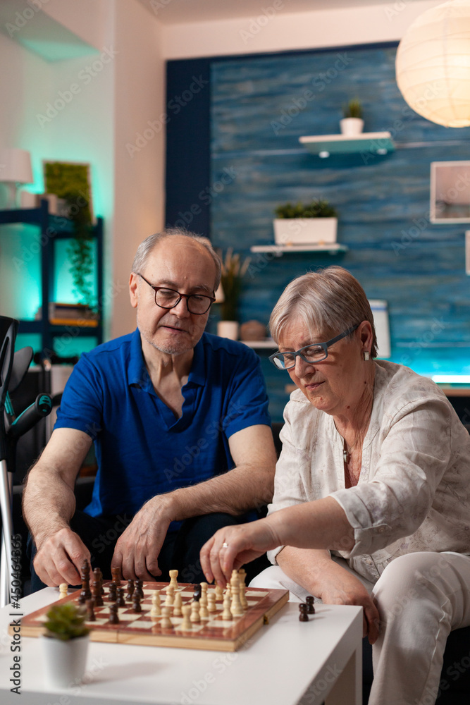 Retirement aged people sitting on living room sofa at home while playing chess game on board to relax. Caucasian old couple enjoying indoor fun activity resting on couch with crutches Retirement aged people sitting on living room sofa at home while playing chess game on board to relax. Caucasian old couple enjoying indoor fun activity resting on couch with crutches