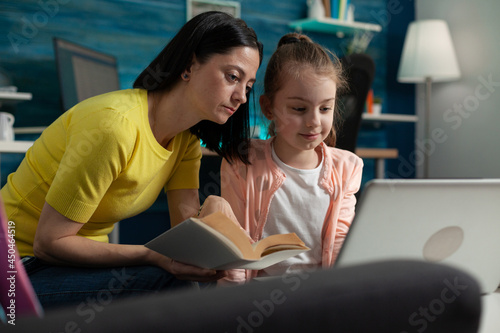 Little pupil using laptop for homework and mom assisting giving help while reading book for online meeting call with classroom and teacher. Caucasian family of two using internet technology