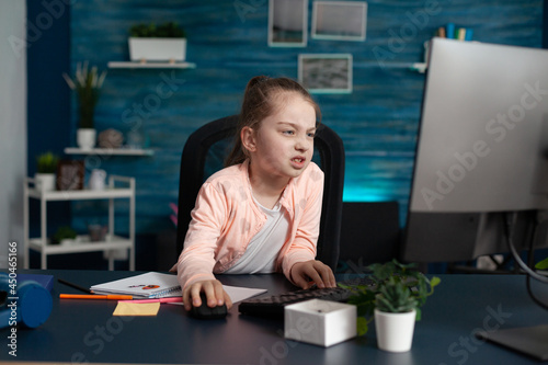 Tired elementary student joining online lesson from home feeling sad about taking notes and doing schoolwork for stressful exam. Little girl being overwhelmed for having school assignments