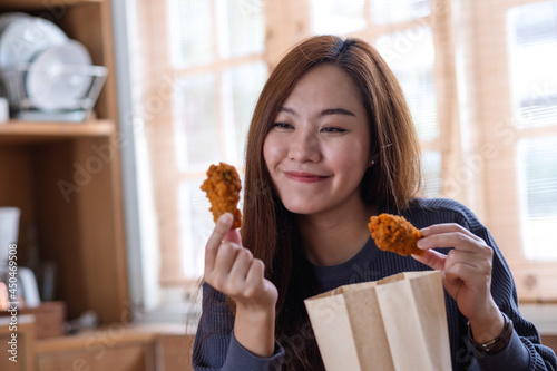 Portrait image of a beautiful young asian woman picking fried chicken from paper food bag to eat at home for food delivery concept