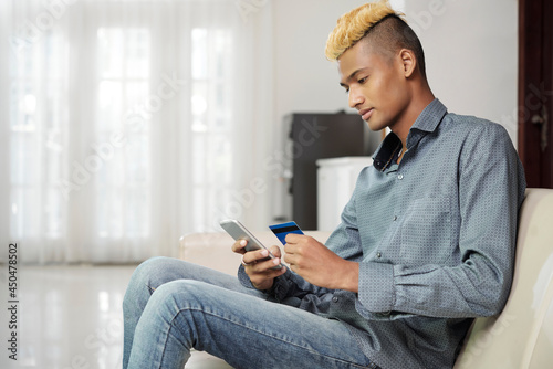 Young man with stylish hairstyle paying with credit card when shopping online via application on smartphone