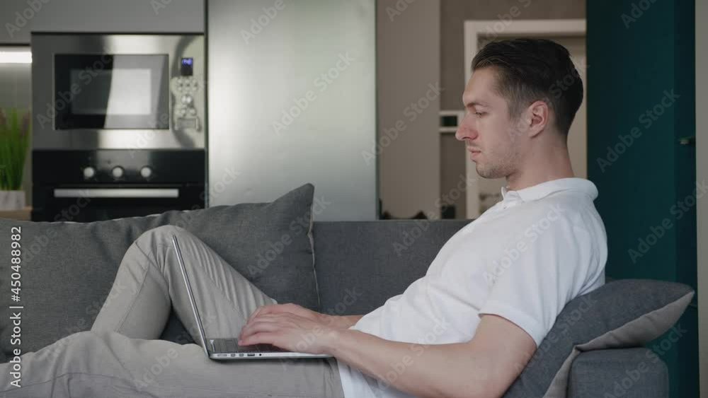 Young man freelancer using laptop device while lying on sofa at home, portrait of male working distantly on computer in apartment Young man freelancer using laptop device while lying on sofa at home, portrait of male working distantly on computer in apartment