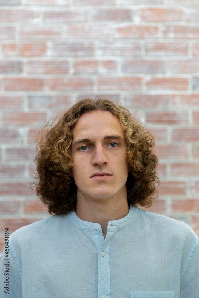 Headshot of young caucasian man looking at camera with blond curly hair. Copy space. Vertical image. Brick wall background.