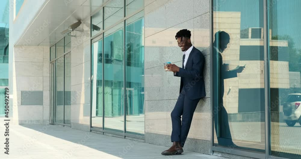 African american businessman drinking coffee and text messaging using mobile phone leaning on wall outside office building African american businessman drinking coffee and text messaging using mobile phone leaning on wall outside office building