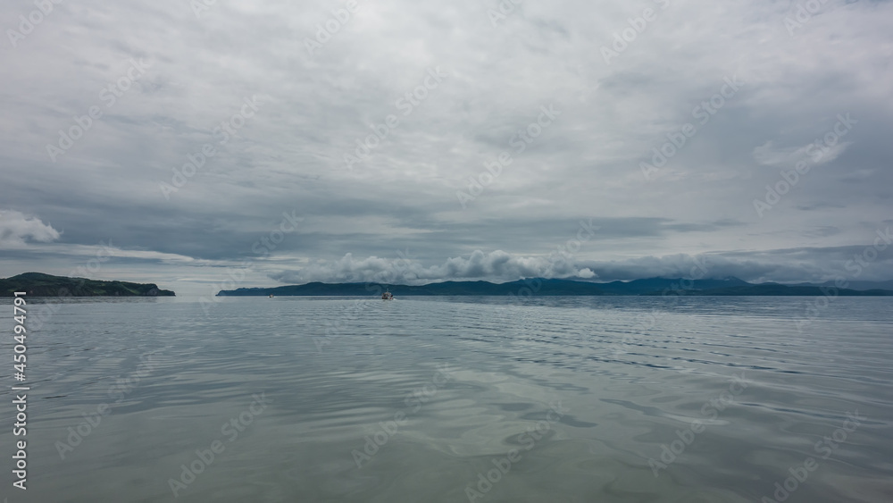 There are light ripples on the surface of the water. A ship is visible in the distance. There is a mountain range on the horizon, hidden in the clouds. Cloudy and quiet. The Pacific Ocean.