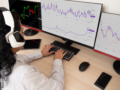 Caucasian young woman sitting at a table doing stock trading on a computer with three black monitors in a white room