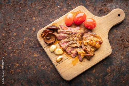 juicy medium rare grilled beef fillet steak meat with tomato, mushroom, garlic and oregano on wooden cutting board on rusty texture background with copy space for text, top view